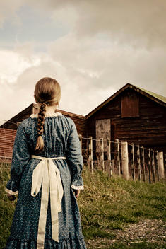 A young girl with her long brown hair in a plait/braid, dressed in an olden day style blue dress with cream bow, stands looking towards a rustic barn. The sky is full of grey and white clouds. Long green grass, and a country fence runs from the girls acro