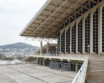White geometric sports venue with blue smoked windows below a large overhanging slanting roof. A low hill rolls through the distant landscape.