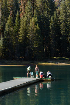 A group of kids (ages 5-12) wearing sun hats and lifejackets engage in summer vacation fun playing at the end of a dock on a scenic lake, trying to catch fish with nets.