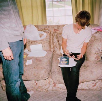 Portrait of a young teenage boy as he counts with his fingers holding his iPhone device and pen in mouth and stack of paper on his leg sitting on the couch with his friend patiently standing nearby in the living of his family\'s suburban home. Pleasant Gro