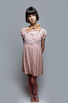 Cute young woman wearing old style pink dress and large bowtie. Short, dark haircut. Neutral background.