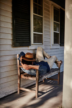 Man laying on bench with hat outside farmhouse