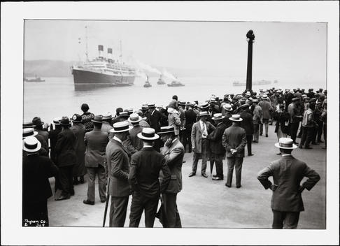 Four Tugboats Escorting An Unidentified Ocean Liner Into A Harbor As A Crowd Watches From The Pier.