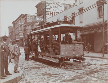 A Street Corner With Trolley Waiting At Center Men Stand On The Corner And Sit On The Trolley And Three Story Businesses Line The Street In The Background, Among Them The Huber Brewery.