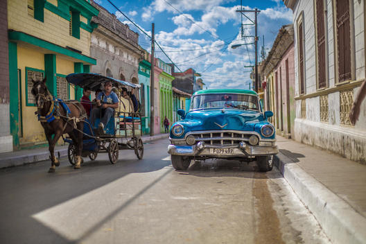 A horse drawn taxi passes a vintage blue car on a colorful street.