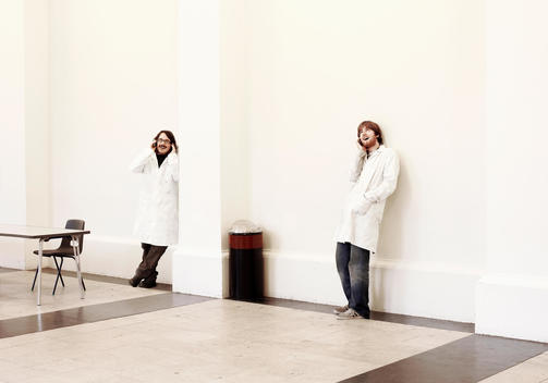 Two Men Leaning Against Wall Wearing Lab Coats And Talking On Their Mobile Phones