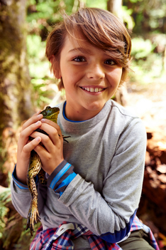 boy seated on a tree trunk holding a green frog in his hand , looking at it , putting over his head smiling and showing to the camera