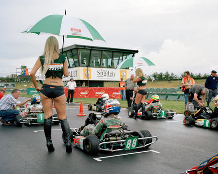 Young Go-Kart Riders Wait On The Starting Grid With Their Support Teams, While Two Blonde Models Hold Umbrellas To Protect Them From The Rain And Help Add A Touch Of Glamour