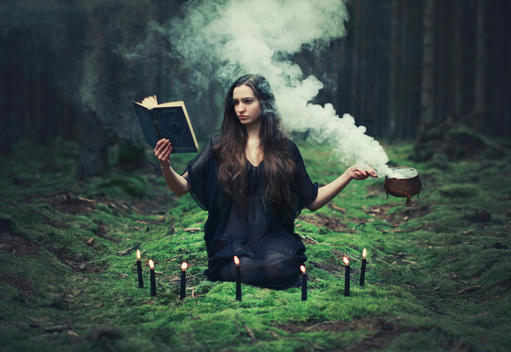 Young adult female with long dark hair wearing a black dress preparing a spell like a witch holding a book and boiling copper cauldron kneeling on green moss surrounded by lit candles in a dark wood