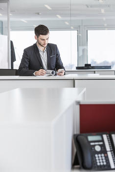 Businessman standing in office reading paperwork and using smartphone