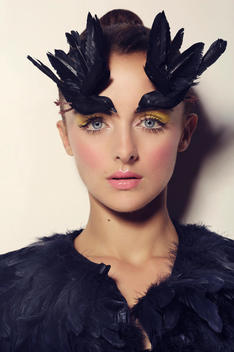 Beauty of a young blue eyes caucasian woman on a white background, with her hair in a bun, wearing 2 black birds in her hair & brows, feather jacket, looking at the lens, yellow eye makeup & glossy lips
