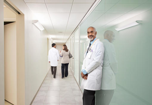 African American male doctor standing in a hospital hallway with a doctor and patient walking in the background.
