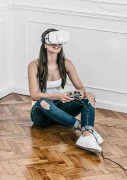 A woman rests on a wooden floor playing a computer game while using a virtual reality headset