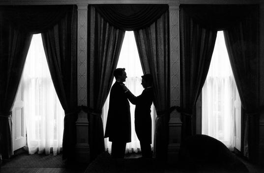 Silhouetted By A Window In A Traditional Hotel Room, The Best Man Ties The Cravat Of The Groom Before The Wedding Ceremony.