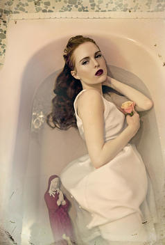 Young woman alone with long dark hair wearing white dress holding a rose lying in a bath of water looking at camera