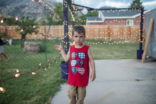 Young boy named Judah runs his hand along a lit chicken wire fence at sunset on a summer evening with the Wasatch mountains at his family\'s suburban home. Pleasant Grove, Utah