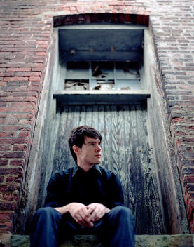 Young Man Seated In Boarded Window, Looking To His Left With His Hands Folded.