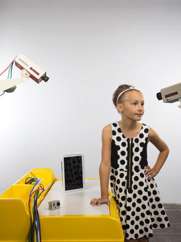A girl in a polka dotted dress stands up next to her desk and looks directly at one of the two surveillance cameras that are pointed towards her