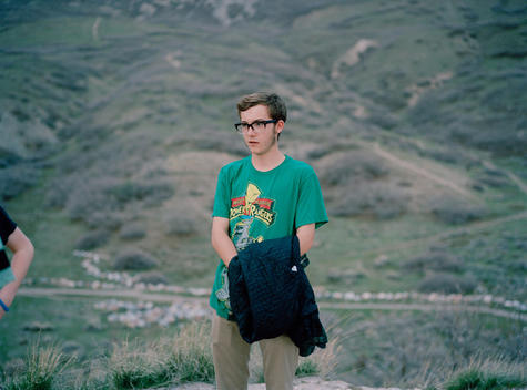 Young nerdy teenage boy wearing a green Power Rangers T-Shirt and thick glasses stands on the top of Mary\'s Hill with friends in the foothills of the Wasatch Mountains behind his family\'s suburban neighborhood. Pleasant Grove, Utah