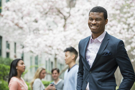 City life in spring. Young people outdoors in a city park. A man in a suit, smiling. Four people in the background. Using cell phones.