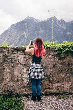 14 year old girl with pink highlights in hair taking photo of mountains in front of wall.