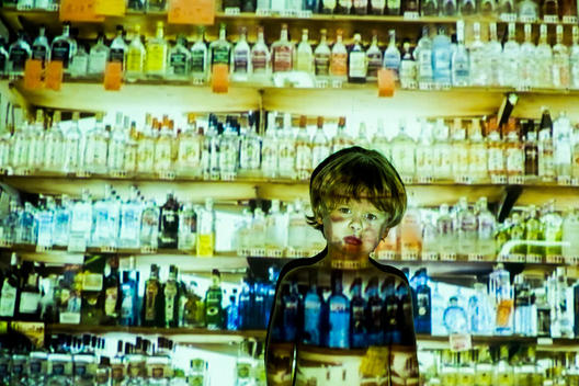 Three and a half year old blonde boy looking at the camera with a projection of liquor shelves on him and the background.