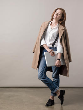 Red headed woman stood in a photography studio with a jacket over her shoulders holding a laptop.