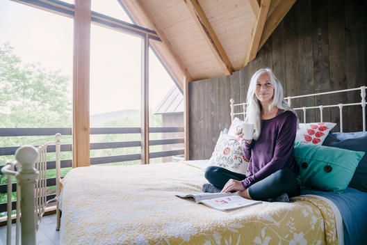 Fit older woman with silver gray hair drinking coffee in bed in a screened in room wearing yoga clothes.