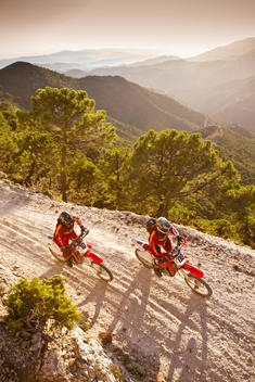 Two off-road motorcycle riders in red riding left to right on dusty trail with mountains in background
