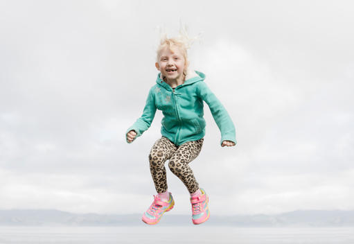 Young white blonde girl jumping and laughing in cheetah pants and pink shoes on overcast day with clouds in background