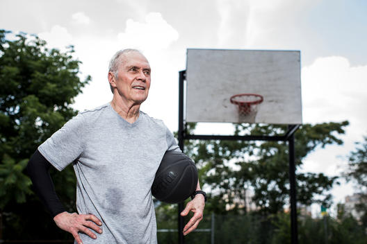 Portrait of an older 50-plus man playing basketball outside on an asphalt court in an urban landscape.