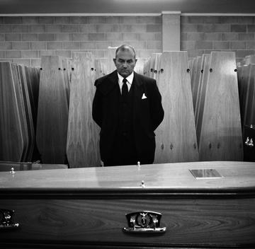 A Male Pall Bearer, Dressed In Black Suit And Tie, At T.Cribb & Sons Funeral Company In East London Stands In Front Of A Coffin Ready To Delivery It To A Local Church For The Funeral Service.