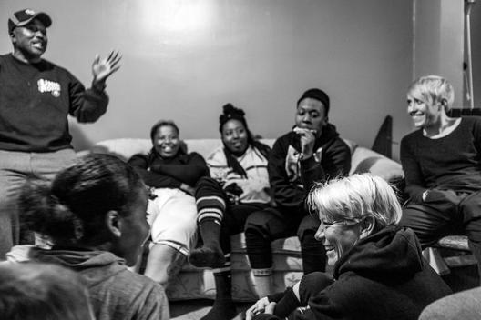 A woman's football team gathers on the couch and floor of their coaches home for a team meeting and to watch game film.