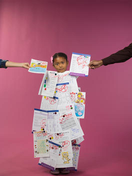 A girl stands covered with graded tests as two more arms are outstretched with more tests