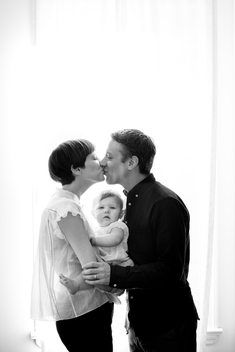 Black and White silhouette image of mom, dad and baby girl kissing in bright light of bedroom window