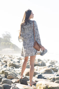 Backlit image of 18 year old brunette girl with long hair in summer dress walking over rocks on rocky beach with purse, flip flops in hand and looking out towards the ocean.