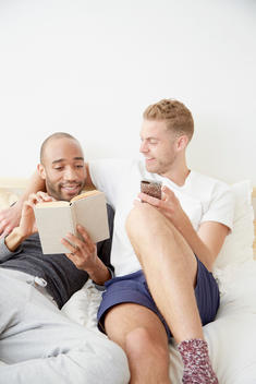 Homosexual couple lying in bed reading book holding smartphone