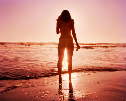 A near silhouette shot from behind of a young woman in a bikini standing at the waters edge on a magenta colored beach.