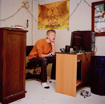 A young shy, introverted teenage boy sits at a small desk in his bedroom where he spends countless hours on the computer browsing the internet