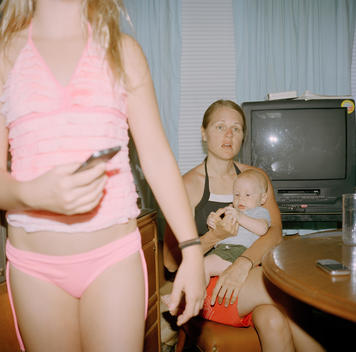 A young mother holds her infant son in the kitchen with her niece wearing a pink swimsuit standing nearby after spending time at nearby Lake Tyler and the family\'s lake house while on vacation. Tyler, Texas