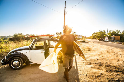 male walking to beach from his VW bug with surfboards under arms