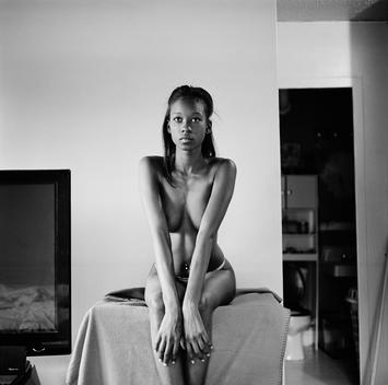 Dramatic black and white portrait of Adia, a beautiful young semi-nude African-American model with navel piercing who lives in the projects/public housing sitting on a box in thong lingerie in her living room. Bronx, NYC