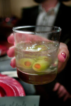 A Woman’S Nail-Polished Hand Holding A Martini At A Cocktail Party That Contains Two Eyeball Like Green Pimento Stuffed Olives.