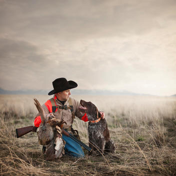 Pheasant hunter wearing cowboy hat kneeling in a field with his double barrel shotgun, petting his dog, and holding his rooster pheasants that he bagged