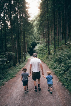 60 something man holding 6 year old and 2 year old boys hands walking down gravel road through forest in Austrian alps.