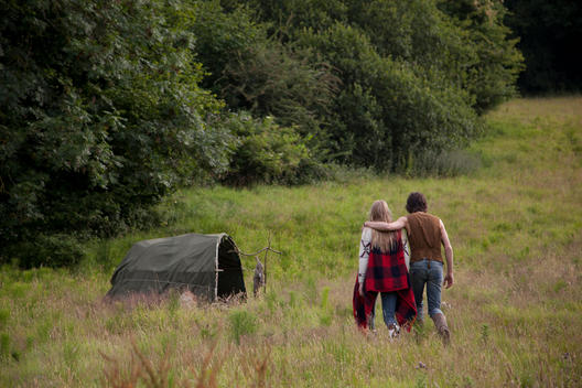 A man and a blonde woman are walking through a field of wild growing grass. They are holding hands, walking towards a tent.