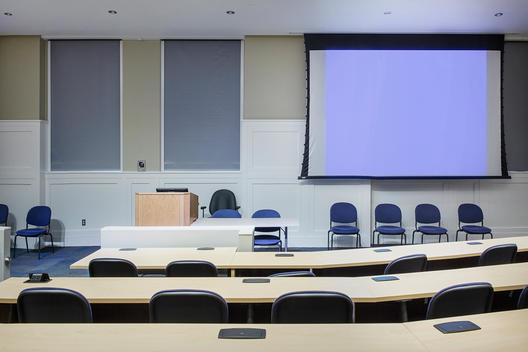 Auditorium style, lecture class room with rows of chairs and long wood tables with a projector screen.