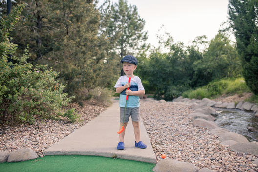 Grant, a young red-head boy, holds his golf club standing alone on the put-put miniature course with a stream nearby as he plays the game with his family. Denver, Colorado