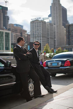 Two Chauffeurs Lean Against A Dark Tinted Car On A Chicago Street.