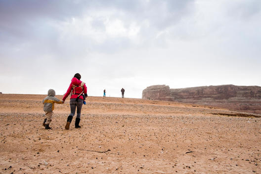 mother in red jacket with baby in a pack and 4 year old son walking in very arid looking area by lake Powell in Arizona with father and son in the background.